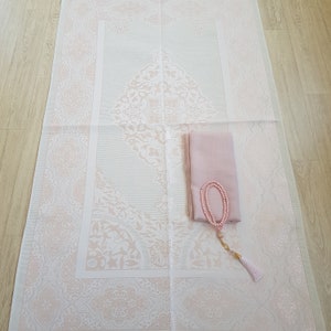May include: A white prayer rug with a pink floral design. A pink scarf and a pink prayer bead necklace are placed on the rug.
