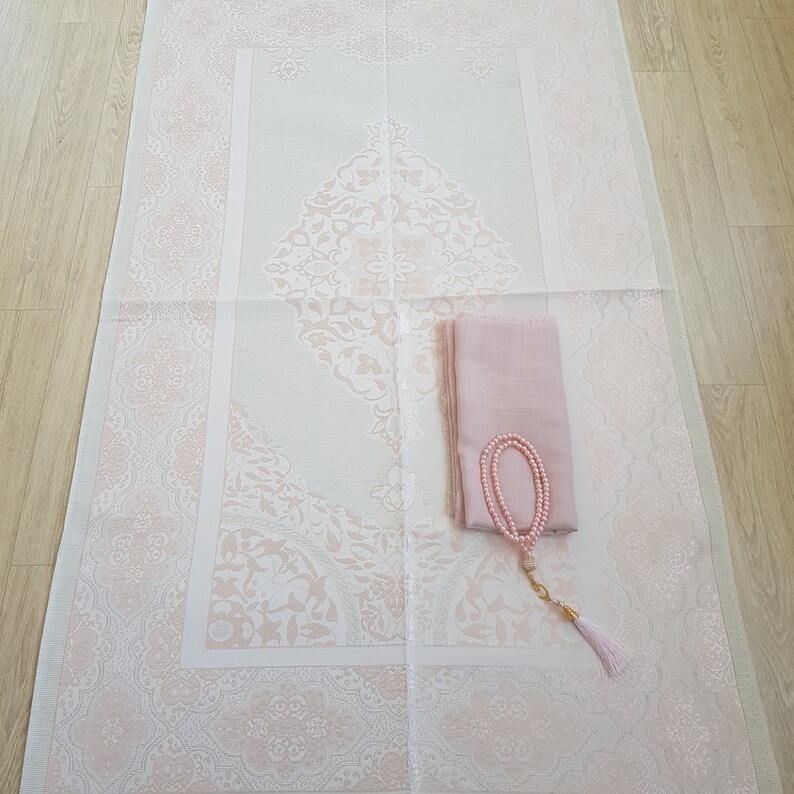May include: A white prayer rug with a pink floral design. A pink scarf and a pink prayer bead necklace are placed on the rug.