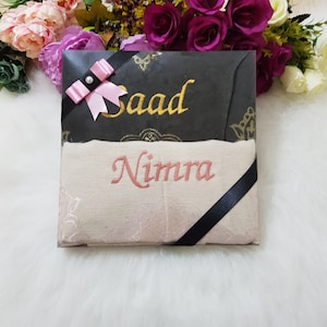 Puede incluir: Una caja de regalo blanca con una cinta negra y un lazo rosa. La caja está decorada con un diseño dorado y los nombres "Saad" y "Nimra" están bordados en la caja.