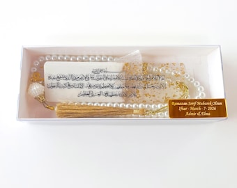 May include: A white beaded prayer beads set with gold accents, presented in a clear box. Includes a card with Arabic script and a gold-colored tag with text. The set is designed for religious use.