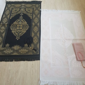 May include: Two prayer rugs, one black with gold accents and one white with pink accents, with prayer beads on top of the white rug.
