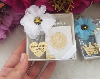 May include: Two white gift boxes with gold accents. The box on the left has a white flower, a heart-shaped tag that says "Sarah & Ali Wedding 10.11.22", and a small white book with a gold inscription. The box on the right has a blue flower, a crown, and a tag that says "Welcome Baby Ali 09.11.22".