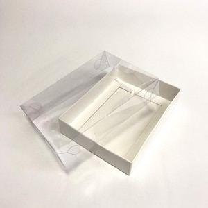 May include: A white rectangular box with a clear plastic lid. The lid is partially open, revealing the interior of the box. The box is empty and sits on a white surface. The box is likely for packaging or storage.