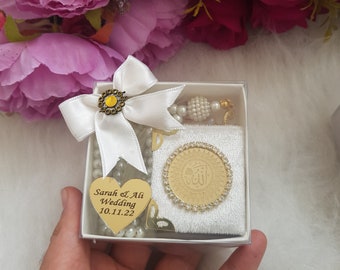 May include: A white gift box with a white bow and a gold heart charm with the text "Sarah & Ali Wedding 10.11.22". The box contains a white prayer rug with a gold Islamic design, a white prayer bead necklace, and a gold charm.