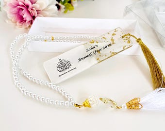 May include: A white pearl necklace with a gold clasp and tassel, paired with a bookmark. The bookmark has gold leaf accents and the text "Soha's Annual Iftar 2026" and a graphic with Arabic text. A white box and flowers are in the background.
