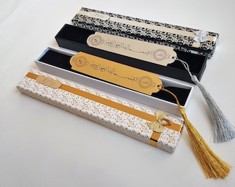 May include: Three gold bookmark boxes with gold tassels. The bookmarks have Arabic calligraphy and are in black boxes with white and black patterned boxes. The boxes are on a white surface.