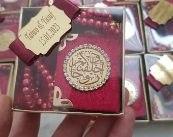 May include: A burgundy velvet prayer book and prayer beads set in a clear box. The book has a gold emblem with Arabic script and a rhinestone border. A gold tag with the names Taiwo & Yusuf and the date 15.01.2023 is attached.