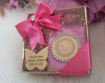 May include: A pink gift box with a pink ribbon and a gold butterfly charm with the text "Welcome Baby Noor 09.11.22". The box also contains a pink velvet pouch with a gold medallion featuring the Arabic word for "Allah" surrounded by rhinestones.