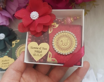 May include: A small white box with a red velvet pouch inside. The pouch has a gold circle with Arabic calligraphy and rhinestones. The box also includes a heart-shaped tag with the text "Jasmine & Yusa Nikkah 02.11.22" and a string of red beads.