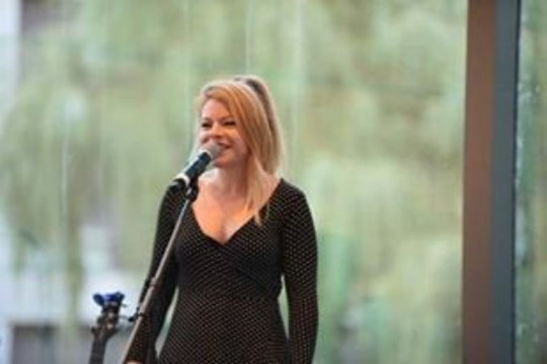 May include: A woman with blonde hair sings into a microphone. She wears a black dress with white polka dots and long sleeves. A microphone stand is in front of her, and a guitar is visible in the background. The setting appears to be indoors.