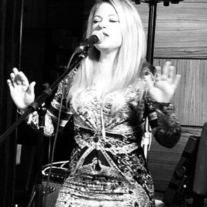 May include: A woman singing into a microphone on a stage. She is wearing a patterned dress with long sleeves. The image is in black and white, with a microphone stand and speaker visible in the background.