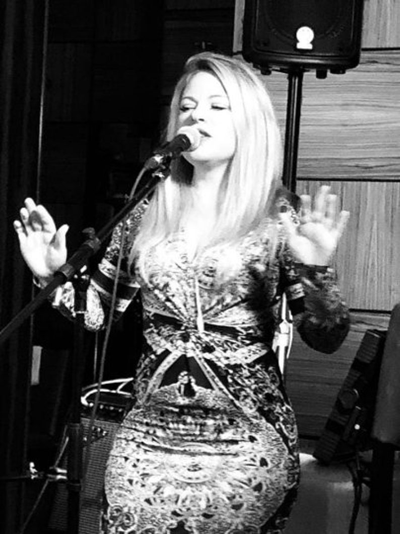 May include: A woman singing into a microphone on a stage. She is wearing a patterned dress with long sleeves. The image is in black and white, with a microphone stand and speaker visible in the background.