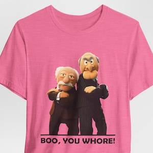 Puede incluir: Camiseta rosa con un gráfico de dos personajes de los Muppets, Statler y Waldorf, con el texto "BOO, YOU WHORE!"
