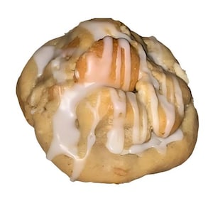 May include: A close-up of a single, round, frosted cookie with a white icing drizzle. The cookie has a light brown color and a soft, chewy texture. The icing is thick and creamy, and it covers most of the cookie.
