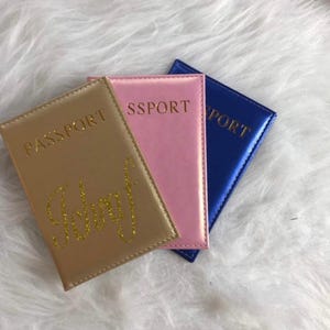 Peut inclure: Trois porte-passeports en or, rose et bleu avec le mot "PASSPORT" imprimé sur chacun. Le porte-passeport doré porte le nom "Jeffrey" écrit en paillettes dorées.