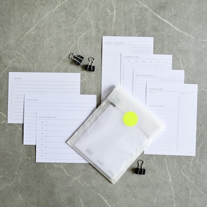 Könnte beinhalten: Eine Sammlung weißer Papiereinlagen für Planer mit verschiedenen Layouts, darunter "To Do", "Notizen" und "Wochenplan". Die Einlagen werden von schwarzen Büroklammern und einer kleinen Verpackung mit einem gelben Aufkleber begleitet, alles auf einer grauen Marmoroberfläche angeordnet.