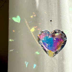 Può includere: Un acchiappasole di cristallo trasparente a forma di cuore è appeso a un filo sottile. Il cristallo rifrange la luce, creando un arcobaleno di colori sulla tenda bianca dietro di esso. L'acchiappasole è sospeso davanti a una tenda bianca.