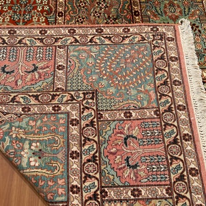 May include: Close-up of a detailed area rug with a floral pattern. The rug features a combination of colors, including teal, pink, and brown. The design includes floral and tree motifs, with a fringed edge.