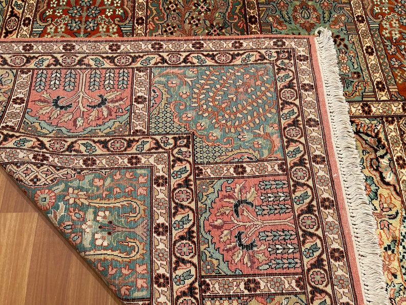 May include: Close-up of a detailed area rug with a floral pattern. The rug features a combination of colors, including teal, pink, and brown. The design includes floral and tree motifs, with a fringed edge.