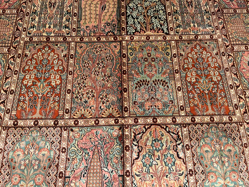 May include: Close-up of a detailed, patterned rug with a repeating design. The rug features a variety of colors, including red, pink, green, and beige. The pattern includes stylized trees and floral motifs, creating a symmetrical and ornate appearance.