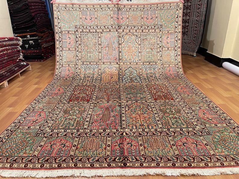 May include: An intricately designed area rug with a repeating pattern of stylized trees, floral motifs, and arched panels in shades of green, red, pink, and beige. The rug has a detailed border and a fringed edge.