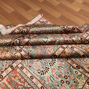 May include: An intricately patterned rug with a folded design. The rug features a complex floral and geometric pattern in shades of teal, peach, cream, and brown. The edges are finished with a pink border and fringe.