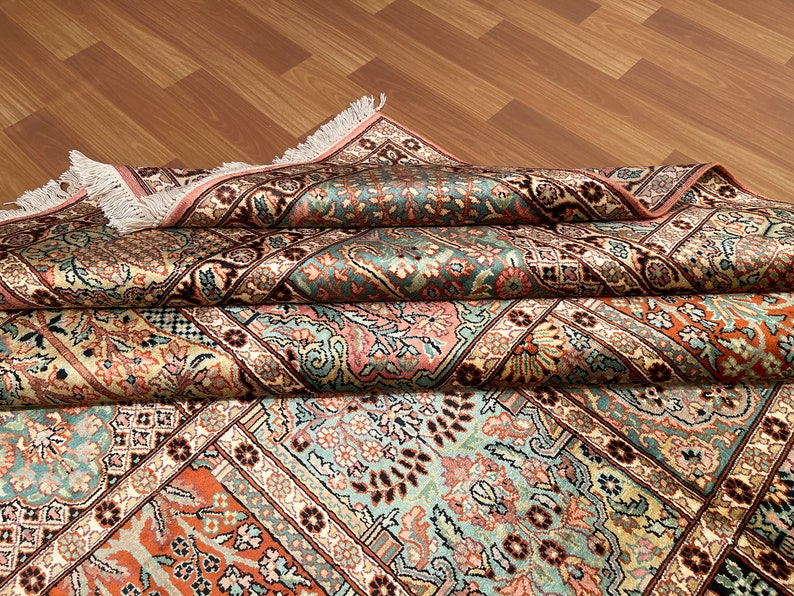 May include: An intricately patterned rug with a folded design. The rug features a complex floral and geometric pattern in shades of teal, peach, cream, and brown. The edges are finished with a pink border and fringe.