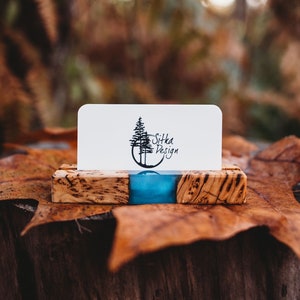Peut inclure: Un porte-cartes de visite en bois avec un incrustation de résine bleue. Le porte-cartes est posé sur une feuille brune. La carte de visite dans le porte-cartes a un logo de deux arbres et le texte "Sitka Design".