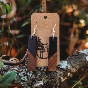 Könnte beinhalten: Holz-Ohrhänger mit geometrischem Design. Jeder Ohrring hat eine rechteckige Form mit einem dunklen Holzoberteil und einem helleren Holzunterteil. Die Ohrringe hängen an silberfarbenen Haken. Im Hintergrund befindet sich ein braunes Etikett mit einem Baumlogo.
