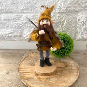 Peut inclure: Figurine de gnome en feutre fait à la main, avec un chapeau jaune avec une plume, une longue barbe brune et une lance. Le gnome porte un manteau jaune, une ceinture marron, un pantalon gris et des bottes noires. La figurine est sur une base en bois et un plateau rond en bois.