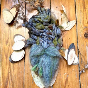 May include: A whimsical felt sculpture of a forest spirit with a green and blue beard, brown eyes, and a mossy head. The sculpture is mounted on a birch wood plaque.