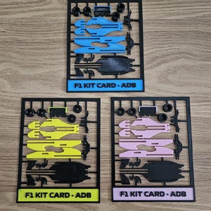 Peut inclure: Trois cartes de kit F1 en bleu, jaune et rose, chacune avec des cadres et des composants noirs. Le texte "F1 KIT CARD - ADB" est imprimé sur chaque carte. Les cartes sont disposées sur une surface en bois.