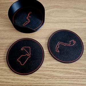 May include: Three black coasters with a red outline of a race track. The coasters are round and have a textured surface.
