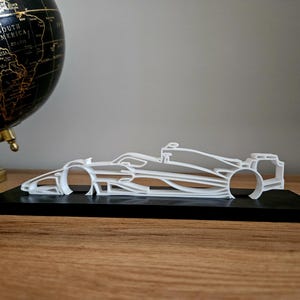 May include: A white, minimalist Formula 1 race car model, displayed on a black base. The car's design is a skeletal outline, showcasing the vehicle's form. The model is set against a backdrop of a globe and a wooden surface.