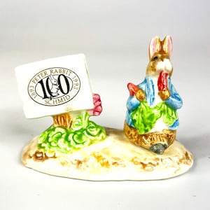 Puede incluir: Una figurita de porcelana de Peter Rabbit sosteniendo una zanahoria y lechuga, de pie sobre una base con un letrero que dice "Peter Rabbit 1893 100 1993 Schmid".