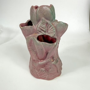 May include: A handcrafted, antique-style vase in a muted mauve colour with green accents. The vase is shaped like a stylized flower with multiple openings and detailed leaf patterns. The interior is a deep red.