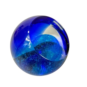 Puede incluir: Un pisapapeles de vidrio redondo, con un exterior azul oscuro y capas internas de azul claro y turquesa. El orbe transparente tiene una superficie lisa y pulida y un diseño único y artístico. Un artículo decorativo.