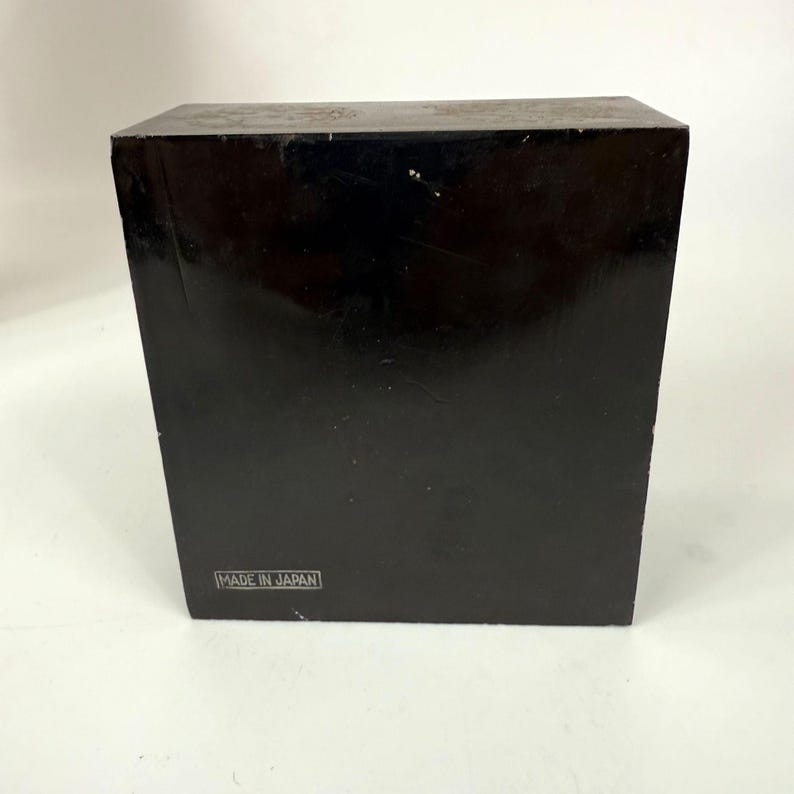 May include: A black, rectangular box with a glossy finish. The box has a label that reads "MADE IN JAPAN" in a white, rectangular box. The box is sitting on a white surface.