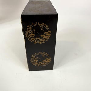 May include: A tall, rectangular black box with gold floral designs. The box has a hinged lid and is decorated with two circular patterns of stylized flowers and foliage. The box appears to be made of wood and has a slightly worn appearance.