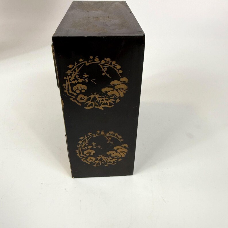 May include: A tall, rectangular black box with gold floral designs. The box has a hinged lid and is decorated with two circular patterns of stylized flowers and foliage. The box appears to be made of wood and has a slightly worn appearance.