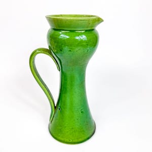 May include: A handmade, green ceramic pitcher with a curved handle and a spout. The pitcher has a glossy finish and a tall, slender shape. The pitcher is approximately 9 inches tall.
