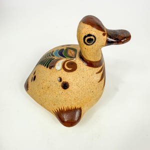 May include: A ceramic duck figurine with a speckled tan body, brown beak, and head. The duck has decorative painted details in blue, green, and brown. The figurine has a rustic, handcrafted appearance.