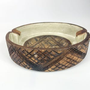 Può includere: Un posacenere rotondo in ceramica con una glassa marrone e bianca. Il posacenere presenta un motivo geometrico all'esterno e un interno bianco liscio.