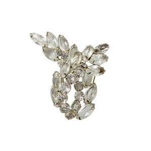 Könnte beinhalten: Vintage Brosche in Silber mit floralem Design. Die Brosche hat klare, oval geformte Strasssteine und kleinere, runde, klare Steine. Die Steine sind in einer Anordnung angeordnet, wodurch ein funkelndes, elegantes Accessoire entsteht.