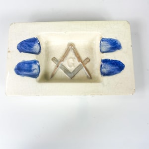 Könnte beinhalten: Ein weißer Aschenbecher aus Keramik mit einem blauen und grauen Freimaurersymbol in der Mitte. Der Aschenbecher hat vier blaue Akzente an den Rändern.
