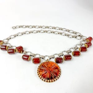 May include: A vintage necklace featuring a silver-tone chain with red barrel beads and a large, round pendant. The pendant has a gold-tone frame and a detailed, reddish-orange design. The necklace is displayed against a white background.