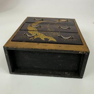 May include: A small, three-drawer wooden chest with a black exterior and gold trim. The drawers are black with gold and yellow bird and floral designs. Each drawer has a small, silver-colored handle.