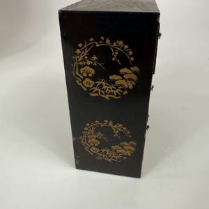 May include: Black wooden cabinet with gold floral designs. The cabinet has a rectangular shape with multiple small drawers. The gold designs are circular and feature flowers, leaves, and other natural elements.