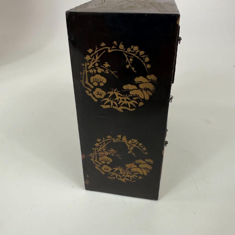 May include: Black wooden cabinet with gold floral designs. The cabinet has a rectangular shape with multiple small drawers. The gold designs are circular and feature flowers, leaves, and other natural elements.