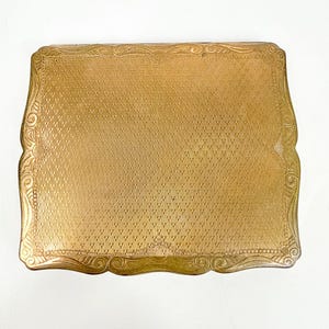 Puede incluir: Un estuche compacto rectangular dorado con una superficie texturizada con trama cruzada. Los bordes están adornados con un diseño ornamental en espiral. El estuche es vintage y para cosméticos.